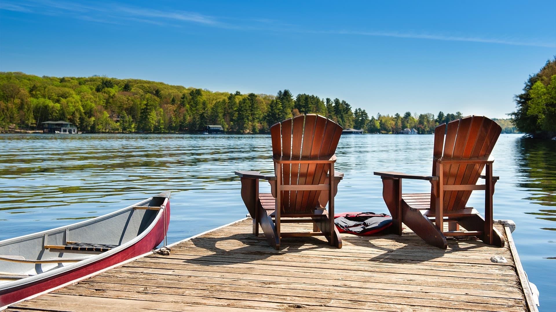 A scenic lakeside view from a dock with two Muskoka chairs and a canoe.